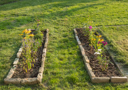 Two rectangle flower bed with sandstone stone border with colorful blooming flowers in a country garden grassの写真素材