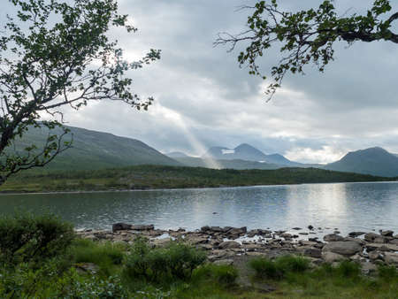 rocky shore of Tarra river. Tarraatno with green hills, birch tree forest and mountains at Padjelantaleden hiking trail. Summer cloudy evening at Lapland, Swedenの写真素材