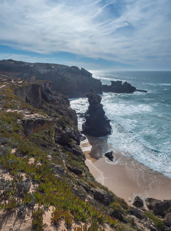 View of sharp cliffs and small sand beach with ocean waves, rocks, stones and green vegetation at wild Rota Vicentina coast near Vila Nova de Milfontes, Portugal.の写真素材