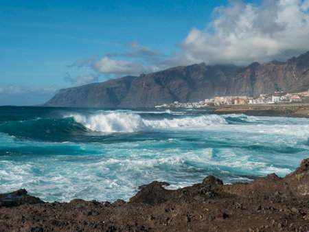 Big breaking waves splash against black lava rock coast. Turquoise blue Atlantic ocean with view of Los Gigantes cliffs and village. Tenerife, Canary islands, Spain. Sunny winter dayの写真素材