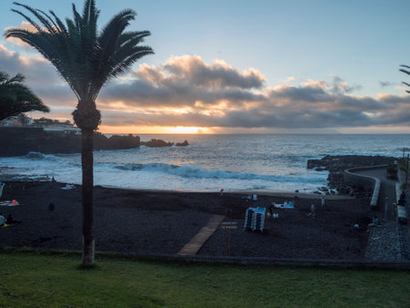 Santiago del Teide, Tenerife, Canary Islands, Spain, December 21, 2021: Sunset view of black sand beach Playa de la Arena with dramatic orange clouds and relaxing tourist people and palm treesの写真素材