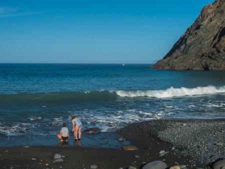 Two woman and child from back at Playa de Vallehermoso, volcanic black sand beach with pebbles and mountain cliff with blue ocean and sky. Vallehermoso, La Gomera, Canary Islands, Spainの写真素材