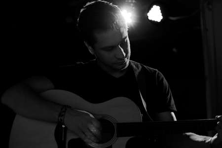 A young handsome guy of European appearance plays an acoustic guitar and sits on a chair. Black and...