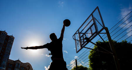 Silhouette of a man who throws a ball into a basketball hoop on the street against a sky with clouds in black and white colorの写真素材