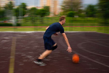 Young redhead guy running and dribbling a basketball at high speed on a sports field on the street during the day motion blurredの写真素材