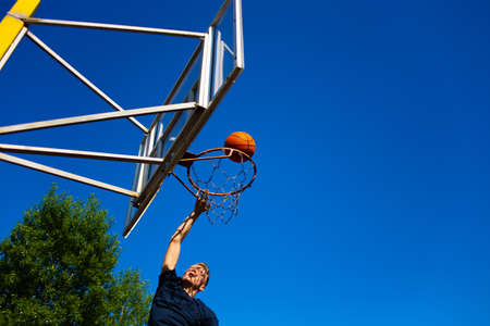 Young red-haired guy in a dark blue t-shirt throws a ball in motion into a basketball hoop against a blue sky outdoorの写真素材