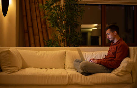 A young man with a beard in a red shirt works with a laptop and sits on the couch in the evening in the house.の写真素材