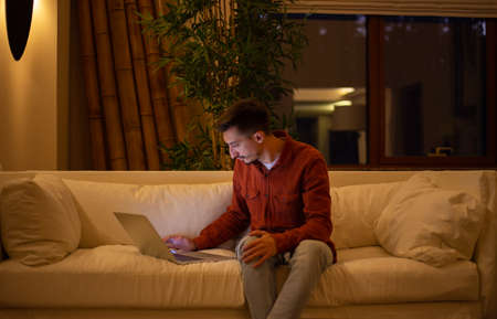 A young man with a beard in a red shirt works with a laptop and sits on the couch in the evening in the house.の写真素材