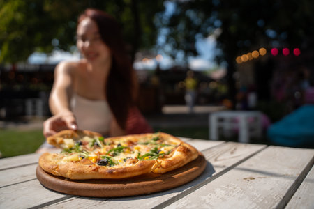 Focused foreground of a hand holding a pizza slice with gourmet toppings, with a blurry background featuring a woman dining outdoors in a lively park with string lightsの写真素材