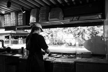 Monochrome image of a pizza chef preparing a dish in an outdoor food stall, with stacked pizza boxes overhead.の写真素材