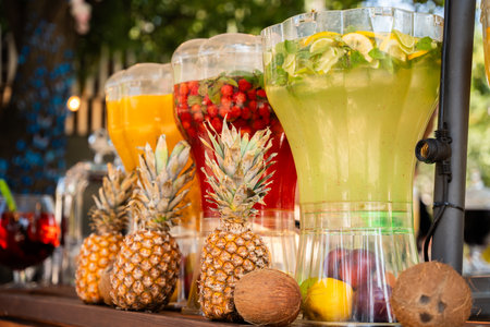 A series of colorful, fruit-infused water dispensers, flanked by pineapples and coconuts, creating a tropical refreshment sceneの写真素材