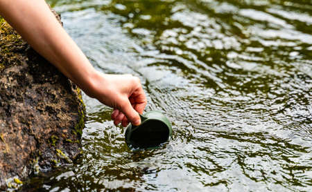 Hand holding a hikers folding cup, reaching to the surface of streaming water to fill the cup.の写真素材