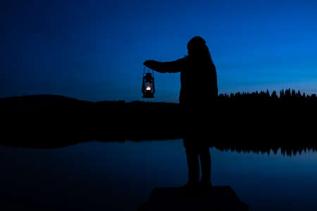 Silhouette of a woman looking into the last light of the day by a lake in the wild, holding a lit vintage kerosene lamp. . High quality photoの写真素材