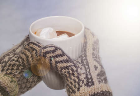 Hands in knitted mittens holding a cup of hot chocolate with marshmallows in a snowy environment. . High quality photoの写真素材