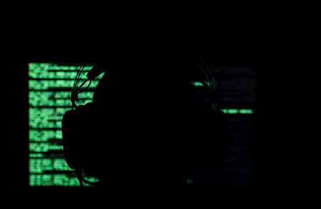 Silhouette of a hacker sitting in front of a monitor with green code in a dark room. Shallow depth of field. High quality photoの写真素材