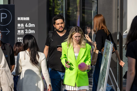 Tokyo, Japan, November 4, 2023: Woman in a Bright Green Blazer Using Smartphone on Busy Street