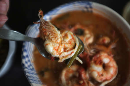Close up Woman hand hold a spoon with shrimp from Tom Yum Goong Spicy Sour Soup or River prawn spicy soup on wood table . Thai foodの写真素材