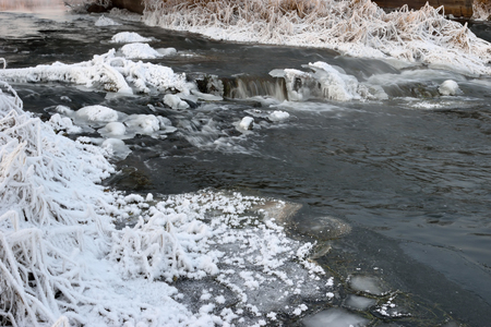 Foaming rapids of the river, covered with growths of ice and bushes dry grass in hoarfrost crystals on the shores of a winter eveningの写真素材