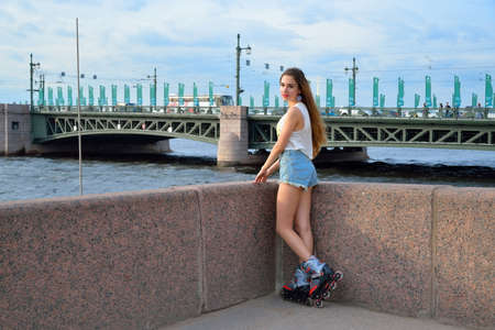 A beautiful girl in roller skates stands on the Neva river embankment against the Palace bridge in...