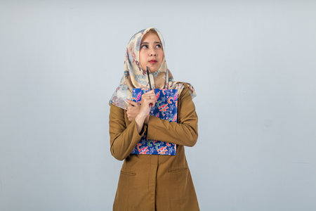 Young Indonesian Woman government employee wearing a brown uniform with thinking face and holding a large bookの写真素材