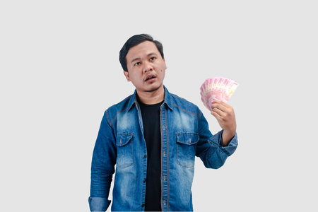 Young Asian man giving love symbol while holding paper money and showing happy expression