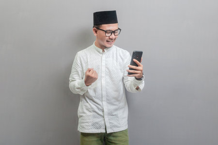 Portrait of an Asian Muslim man wearing a koko shirt and peci with shades of the fasting month, clenched fist showing excitement when looking to his smartphone, isolated on a gray backgroundの写真素材