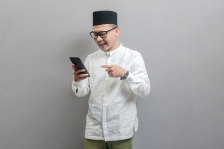Portrait of an Asian Muslim man wearing a koko shirt and peci with shades of the fasting month, smiling pointing on his smartphone, isolated on a gray backgroundの写真素材