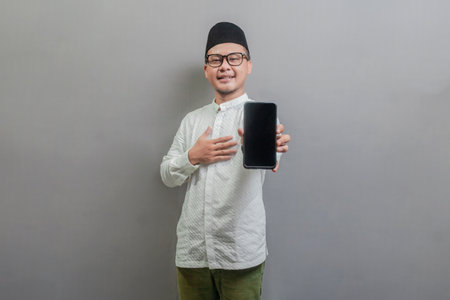 Portrait of an Asian Muslim man wearing a koko shirt and peci with shades of the fasting month, while stroking his chest and showing his smartphone, isolated on a gray backgroundの写真素材