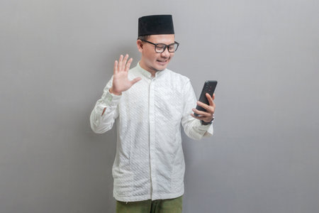 Portrait of an Asian Muslim man wearing a koko shirt and peci with shades of the fasting month, smiling weaving hand on his smartphone, isolated on a gray backgroundの写真素材
