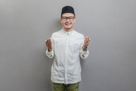Asian Muslim man wearing a koko shirt and peci with shades of the fasting month, standing looking camera happily to show grateful , isolated on a gray backgroundの写真素材