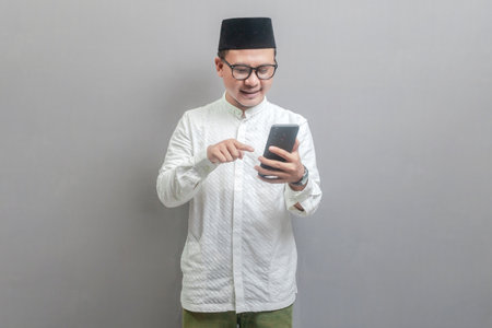 Portrait of an Asian Muslim man wearing a koko shirt and peci with shades of the fasting month, smiling pointing finger on his smartphone, isolated on a gray backgroundの写真素材
