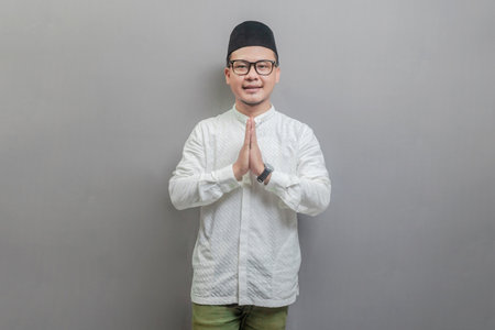Asian Muslim man wearing a koko shirt and peci with shades of the fasting month, standing with Eid greeting gesture and welcoming Ramadan, isolated on a gray backgroundの写真素材