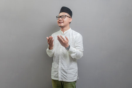 Asian Muslim man wearing a koko shirt and peci with shades of the fasting month, standing looking empty space happily to show grateful , isolated on a gray backgroundの写真素材