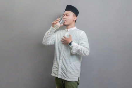 Happy asian muslim man drinking a fresh glass of pure water wearing a koko shirt and peci with shades of the fasting month, isolated on a gray backgroundの写真素材
