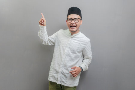 Asian Muslim man wearing a koko shirt and peci with shades of the fasting month, standing looking camera and pointing finger to empty space , isolated on a gray backgroundの写真素材