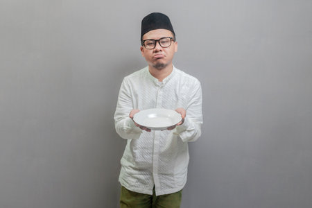 Asian Muslim man wearing a koko shirt and peci with shades of the fasting month, holding a plate while waiting for the time to break the fast, isolated on a gray backgroundの写真素材