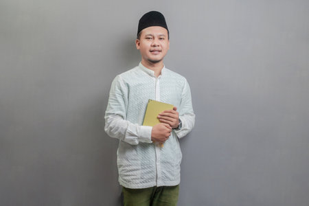 Asian Muslim man wearing a koko shirt and peci with shades of the fasting month, holding prayer beads and reading Qur'an, isolated on a gray backgroundの写真素材