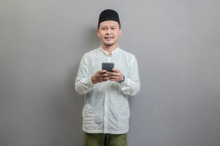 Happy asian muslim man using smart phone cellphone for calls, social media, mobile application online, wearing a koko shirt and peci with shades of the fasting month, isolated on a gray backgroundの写真素材
