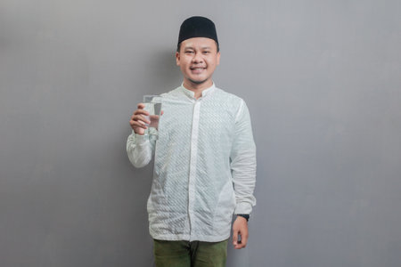 Happy asian muslim man holding glass of pure water give to camera wearing a koko shirt and peci with shades of the fasting month, isolated on a gray backgroundの写真素材