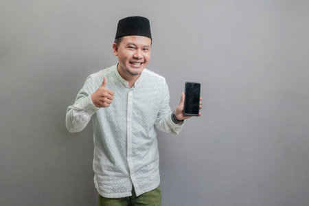 Portrait of an Asian Muslim man wearing a koko shirt and peci with shades of the fasting month, smiling with showing thumbs up and his smartphone, isolated on a gray backgroundの写真素材