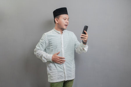 Asian Muslim man wearing a koko shirt and peci with shades of the fasting month, holding back hunger and waiting for the time to break the fast, isolated on a gray backgroundの写真素材