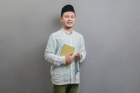 Asian Muslim man wearing a koko shirt and peci with shades of the fasting month, holding prayer beads and reading Qur'an, isolated on a gray backgroundの写真素材