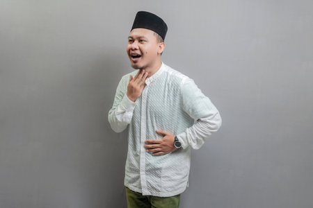 Asian Muslim man wearing a koko shirt and peci with shades of the fasting month, holding back hunger and waiting for the time to break the fast, isolated on a gray backgroundの写真素材