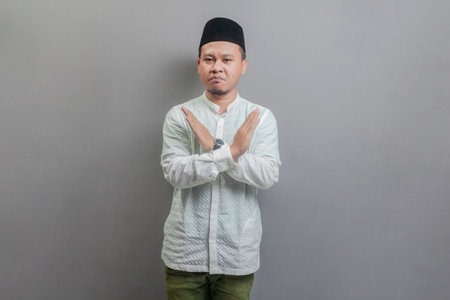Asian muslim man doing stop sign with cross of the hand wearing a koko shirt and peci with shades of the fasting month, isolated on a gray backgroundの写真素材