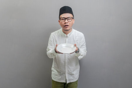 Asian Muslim man wearing a koko shirt and peci with shades of the fasting month, holding a plate while waiting for the time to break the fast, isolated on a gray backgroundの写真素材