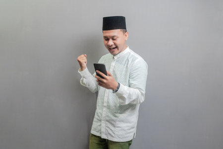 Asian Muslim man wearing a koko shirt and peci with shades of the fasting month, clenching his fist and holding his smartphone, isolated on a gray backgroundの写真素材