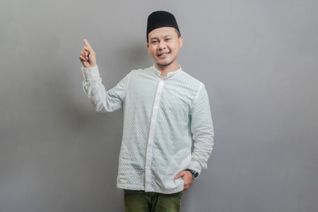 Happy asian muslim man recommend promo as pointing to copy space, indicate down link wearing a koko shirt and peci with shades of the fasting month, isolated on a gray backgroundの写真素材