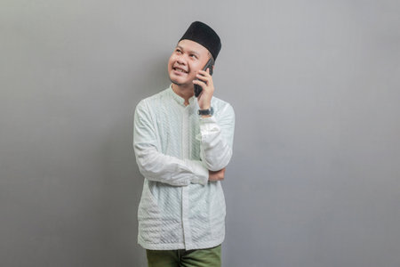 Portrait of an Asian Muslim man wearing a koko shirt and peci with shades of the fasting month, calling with familly or friend, isolated on a gray backgroundの写真素材