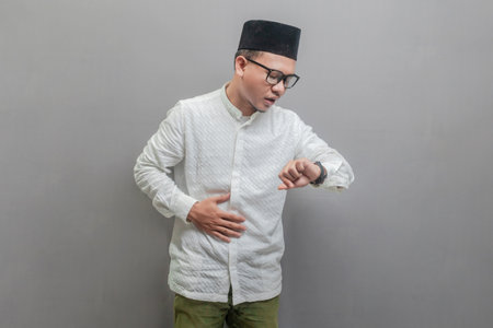 Asian Muslim man wearing a koko shirt and peci with shades of the fasting month, holding back hunger and waiting for the time to break the fast, isolated on a gray backgroundの写真素材