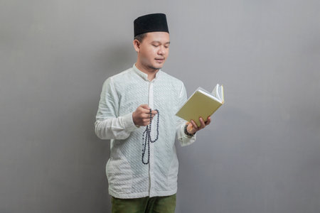 Asian Muslim man wearing a koko shirt and peci with shades of the fasting month, holding prayer beads and reading Qur'an, isolated on a gray backgroundの写真素材